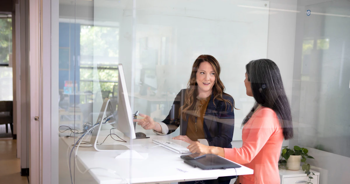 Zwei Frauen, die an einem Stehschreibtisch in einem Büro mit Glaswänden sprechen
